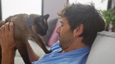 Mavi gömlekli sakallı bir adam, sıcak bir oturma odasındaki kanepede kahverengi siyam kedisiyle etkileşime geçiyor..