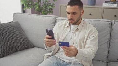 Modern bir oturma odasındaki koltukta kredi kartı ve akıllı telefon tutan odaklı bir İspanyol..