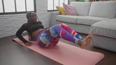 Afro-Amerikalı bir kadın oturma odasında pembe bir paspas, şık taytlar ve örgülerle yoga yapıyor..