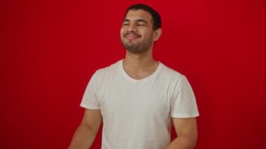 Beyaz tişört giyen genç İspanyol adam gülümserken ve kameraya bakarken yan tarafı işaret ederken elleri ve parmaklarıyla izole edilmiş kırmızı arka planı izliyor.