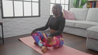 Örgülü Afrikalı bir Amerikalı kadın modern bir oturma odasındaki yoga minderinde meditasyon yapıyor.