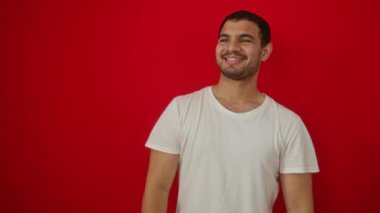 Beyaz tişörtlü genç İspanyol adam yüzünde gülümsemeyle yan yana bakıyor, doğal bir ifade. Kendinden emin gül. izole edilmiş kırmızı arkaplan