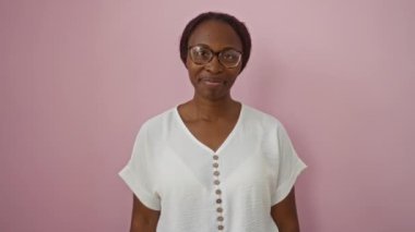 Gözlüklü ve tişörtlü Afro-Amerikalı genç bir kadın, telefonla el hareketi yapıyor ve pembe arka planda izole edilmiş iletişim ve pozitiflik kavramına gülümsüyor.