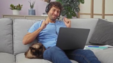 Neşeli bir adam kulaklıkla kulaklıkla müzik dinlemekten hoşlanır ve yanında bir kanepede oturan siyam kedisi..