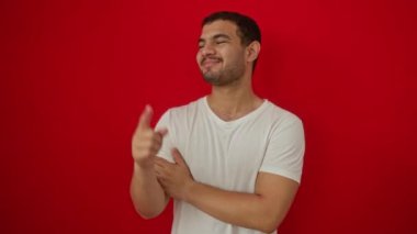 Beyaz tişörtlü, mutlu bir gülümseme, el ve parmak izole edilmiş kırmızı arka planda duran İspanyol genç adam.