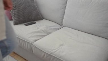 Beyaz gömlek ve kot pantolon giyen genç bir İspanyol modern bir oturma odasındaki gri bir koltukta elinde uzaktan kumandayla oturuyor ve yanında da tekel köpeği var..