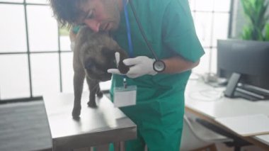 Yeşil önlüklü İspanyol bir adam, parlak ışıklandırılmış bir veteriner kliniğindeki metal bir masada bir siyam kedisini nazikçe muayene ediyor..