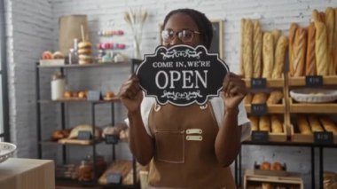 Arkadaki ekmek raflarıyla fırında açık tabela tutan bir kadın.