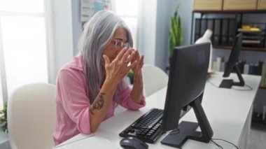 Gri saçlı ve dövmeli yaşlı bir kadın masanın üstünde bilgisayarla ofis ortamında stresli görünüyor.