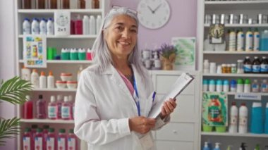 Eczanede elinde bir panoyla duran kadın eczacı elinde bir sürü ürünle gülümsüyor..