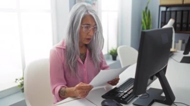 Ofiste bilgisayar başında çalışan gri saçlı kadın, odaklanmış ifadeyle belgeler okuyor.