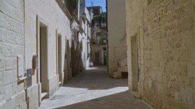 Derste dar, taş döşeli dar bir sokak. Puglia, her iki tarafında kapı ve pencereler olan tarihi mimariyi yakalıyor. Güneş ışığıyla yıkanmış..