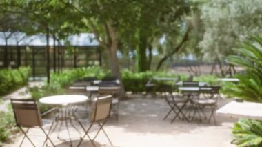 Mallorca 'da bir kafenin ya da restoran terasının bulanık manzarasında ağaçların ve yeşilliklerin altında sayısız boş masa ve sandalye var.