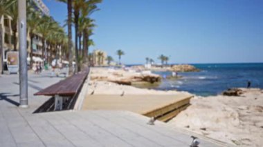 Bulanık bir sahil yolu manzarası. Palmiye ağaçları, banklar ve arka planda gezinen insanlar. Denizin kıyılarla buluştuğu kayalık bir sahil şeridi.