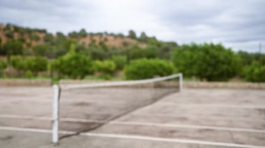 Bulutlu bir günde, arka planda bulanık yeşil yapraklar ve tepelerin olduğu kırsal bir tenis kortunun odaklanmış görüntüsü.