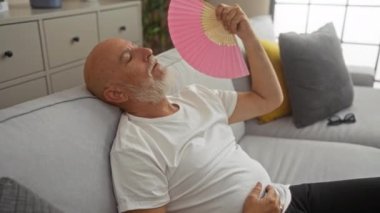 Oturma odasındaki kanepede dinlenen yaşlı bir adam pembe bir yelpazeyle yelpaze takıyor, beyaz bir tişört giyiyor, minderlerle ve ev bitkileriyle çevrili, evde sakin bir anın tadını çıkarıyor.