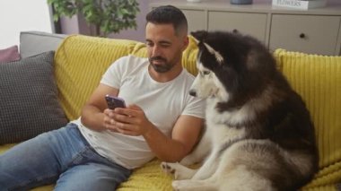 Genç bir İspanyol adam, rahat bir oturma odasındaki akıllı telefonu kullanarak, bir Husky ile koltukta dinleniyor..