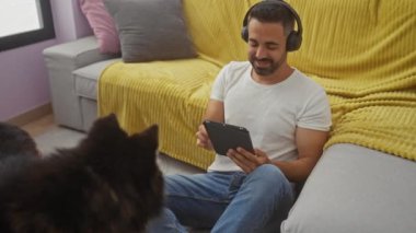 Genç bir İspanyol adam, rahat bir oturma odasında köpeğiyle birlikte tablet ve kulaklıklarıyla vakit geçirir..
