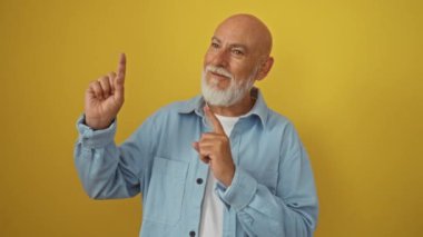 Adam, yaşlı, kel, kot gömlek giyiyor, kenarda duruyor, iki elle işaret ediyor, gülümsüyor ve izole edilmiş sarı arka planda kameraya bakıyor.