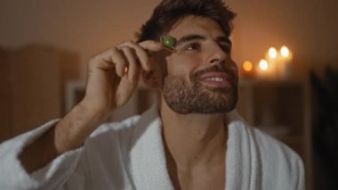 Kaplıca odasında yüzüne yeşim bir silindir takmış genç bir adam arka planda mumlar parıldarken, gözleri kapalı, sakallı ve beyaz bir bornoz giymiş, rahatlatıcı bir anın tadını çıkarıyor.