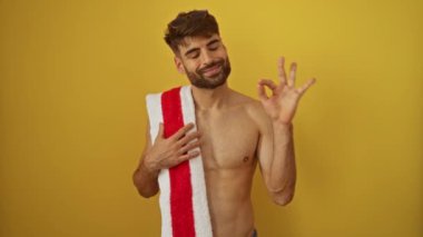 İspanyol asıllı genç adam plaj havlusu tutuyor. Gülümsüyor ve işaret veriyor. Mükemmel bir parmak işareti, dost canlısı ve mutlu.