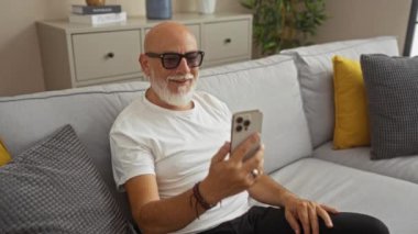 Akıllı telefondaki bir video görüşmesi sırasında gülümseyen bir adam, rahat bir oturma odasında rahat bir koltukta oturuyor, iç mekan bitkileri ve arka planda renkli yastıklar var..