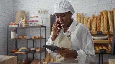 Pastanedeki genç Afrikalı kadın telefonla konuşuyor ve tablet kullanıyor. Arka planda ekmek rafları olan üniforma giyiyor.