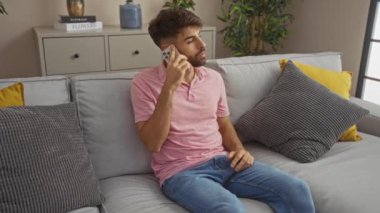 Pembe tişörtlü, sakallı yakışıklı genç bir adam bir kanepede oturuyor ve modern dekorlu rahat bir oturma odasında telefon kullanıyor.