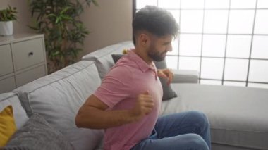 Pembe tişörtlü, sakallı genç adam, modern oturma odasındaki koltukta otururken sırt ağrısı çekiyor. Çiçekler ve yastıklarla..