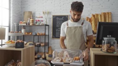 Önlüklü genç sakallı bir adam fırında çalışıyor, arka planda çeşitli ekmek ve kurabiyelerle sergilenen pastaları sergiliyor..