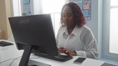 Akıllı telefon kullanarak, iş ortamını ve profesyonel kıyafetleri sergileyerek bilgisayar başında çalışan bir kadın.