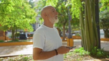 Zengin yeşilliklerle çevrili bir şehir parkında akıllı telefon kullanan bir adam bir kasabada rahat bir açık hava manzarası sergiliyor.