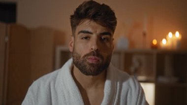 Sağlık merkezinde sakallı, ağzı durulanan, mum ışığında yanan beyaz bir bornoz giyen İspanyol genç bir adam.