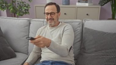 Rahat bir oturma odasındaki koltukta uzaktan kumandayla rahatlayan olgun bir adam.