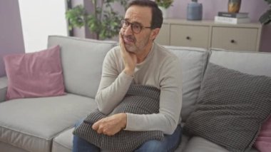 Oturma odasında elinde uzaktan kumandayla oturan düşünceli olgun bir adam.