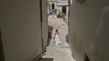 Beyaz elbiseli genç bir İspanyol kadın, eski Ostuni, Puglia, İtalya, Avrupai 'de dar bir sokakta yürüyor. Çiçekleri ve tarihi mimarisi olan büyüleyici bir avluya doğru..