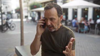Şehir sokağında telefonla konuşan, tişört giyen, sakallı, arka planda açık bir kafe, orta yaşlı, hayal kırıklığına uğramış, halka açık, olgun, konuşan, İspanyol asıllı bir adam..