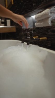 Bir kadın lüks bir banyo ortamında eski bir muslukla köpük banyosu hazırlıyor..