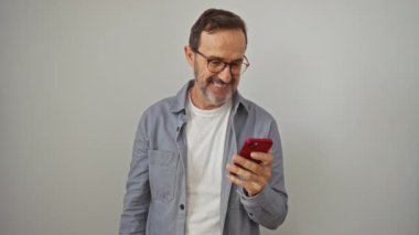 İspanyol bir adam, orta yaşlı, akıllı telefon kullanıyor ve beyaz arka planda izole edilmiş mutlu bir gülümseme ve el hareketleriyle bir tarafa işaret ediyor.