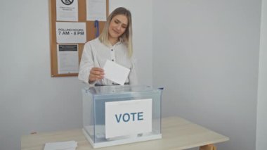 Genç sarışın bir kadın seçim odasında oy veriyor. Gülümsüyor ve onaylıyor. Demokratik süreçten memnun olduğunu ima ediyor..