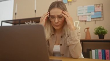 Evde ofis ortamında çalışan sarı saçlı ve gözlüklü genç bir kadın. Dizüstü bilgisayar kullanırken stresli ve odaklanmış görünüyor..