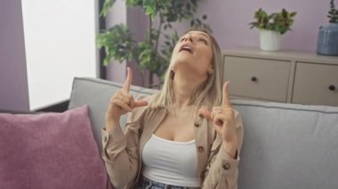 Kanepede oturan genç sarışın kadın şaşırmış bir bakış ve evdeki şaşkın ifadeyle yukarı bakıyor.
