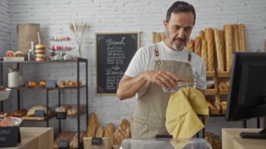 İspanyol pastanede çalışan, önlük giyen, arka planda ekmek ve hamur işleri olan ve duvarda karatahta menüsü olan bir adam.