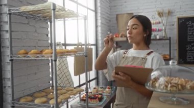 Fırında çalışan genç bir kadın elinde panoyla envanteri kontrol ediyor. Etrafı çeşitli hamur işleri ve tatlılarla çevrili.