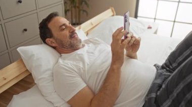 Orta yaşlı bir adam, beyaz yastıklı ve ahşap çerçeveli parlak bir yatak odasında akıllı telefonunu kullanırken evde yatakta dinleniyor.