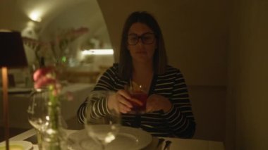 İtalyan restoranında oturan, gözlüklü, limon dilimli, loş bir ortamda içki içen genç bir İspanyol kadın..