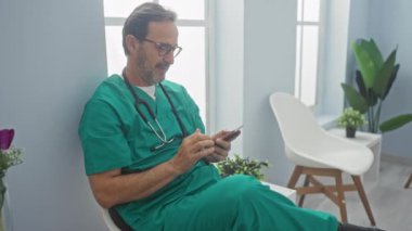 Önlüklü ve gözlüklü yakışıklı doktor klinik bekleme odasında akıllı telefon kullanıyor..