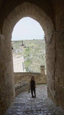 İtalya 'nın tarihi eski kenti Mata' yı keşfeden genç İspanyol kadın, arka planda Avrupa kırsalının manzaralı manzarasıyla taş kemer altında yürüyor..