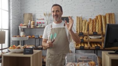 Orta yaşlı bir adam fırın tezgahında çalışıyor. Etrafı ekmek ve hamur işleriyle çevrili. Modern, kırsal bir ortamda neşeli bir tebessümle eşyalar düzenliyor.