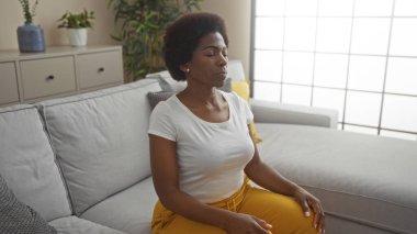 İyi ışıklandırılmış bir oturma odasında, sakinlik ve gevşemeyi vurgulayan beyaz duvarlı gri koltukta meditasyon yapan bir kadın..